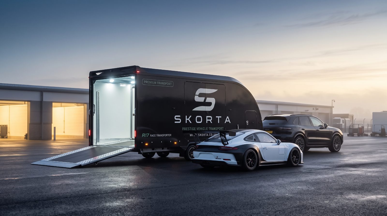 An unbranded GT race car beside the open SKORTA trailer in a dawn paddock
