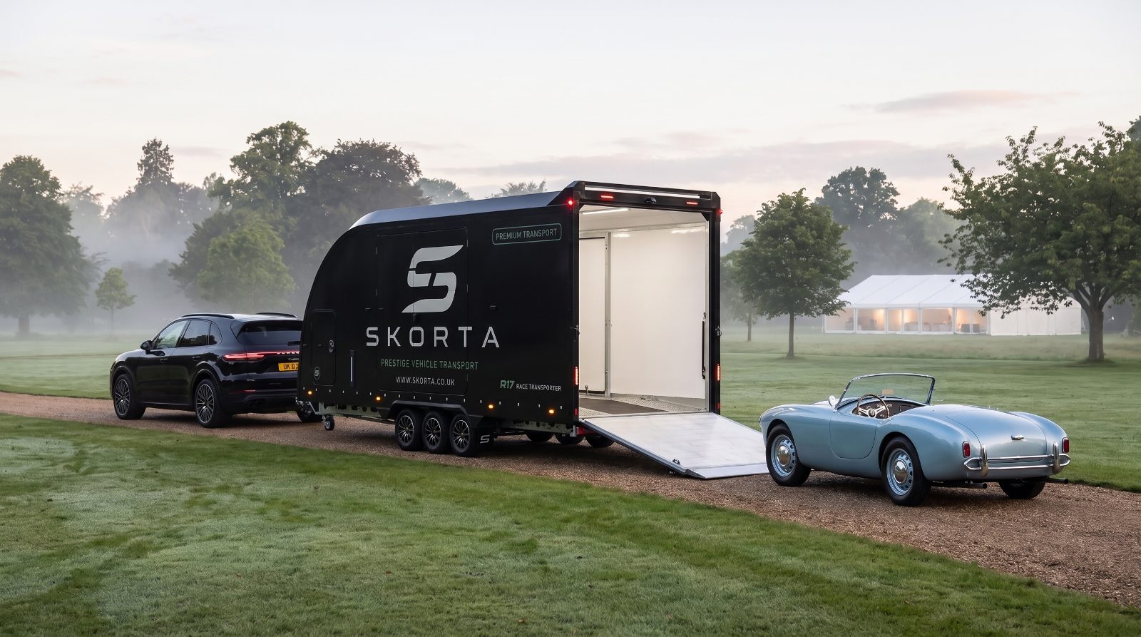 A pale-blue classic roadster beside the open SKORTA trailer on a concours lawn at dawn
